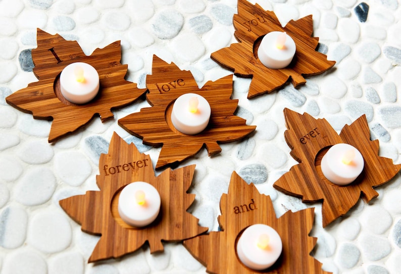 Op de afbeelding: Zes houten kaarshouders in de vorm van esdoornbladeren met de woorden "Ik hou van je voor altijd en eeuwig" erop gegraveerd. Elke houder heeft een klein theelichtje in het midden.