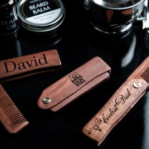 May include: Three wooden pocket combs with personalized engravings. The combs are brown and have a brass screw. The engravings read "David", "World's Coolest Dad", and a logo with the text "The Bearded Balm".