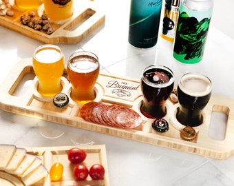 Puede incluir: Una bandeja de vuelo de cerveza de madera con cuatro vasos llenos de diferentes cervezas. La bandeja tiene un asa a cada lado y está grabada con "The Breemont Family" en una fuente decorativa. También hay rebanadas de salami y tapas de botellas en la bandeja.
