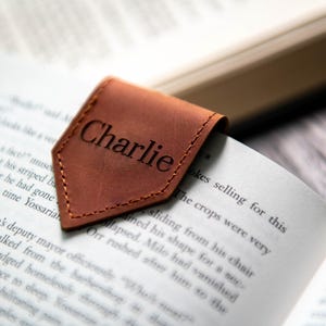 Op de afbeelding: Een bruine leren bladwijzer met de naam "Charlie" erop gegraveerd. De bladwijzer ligt op een boek.