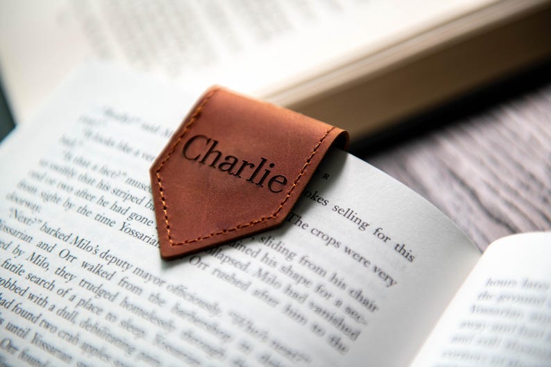Op de afbeelding: Een bruine leren bladwijzer met de naam "Charlie" erop gegraveerd. De bladwijzer ligt op een boek.