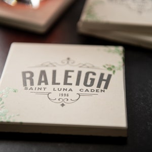 Puede incluir: Un posavasos de cerámica blanca con un diseño en blanco y negro que dice "Raleigh" con los nombres "Saint Luna Caden" y el año "1996" debajo. El posavasos tiene un diseño floral verde en el lateral.