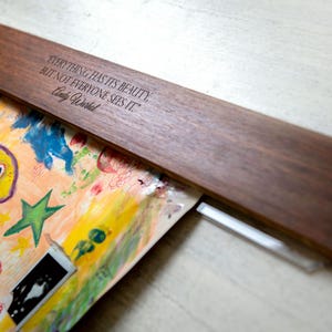 May include: A wooden wall mount with a quote by Andy Warhol: "Everything has its beauty, but not everyone sees it." The mount is holding a colorful abstract painting with a yellow face and stars.