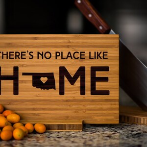 May include: A wooden cutting board with the text "There's no place like home" and an outline of the state of Oklahoma with a heart in the center. The board is sitting on a counter with a knife and some small, orange fruits.