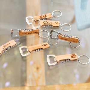 Op de afbeelding: Een set van zeven houten flessenopener sleutelhangers met verschillende gegraveerde spreuken en namen. De sleutelhangers zijn gemaakt van lichtbruin hout en hebben een zilveren flessenopener en kurkentrekker. De sleutelhangers worden op een glazen oppervlak gepresenteerd.