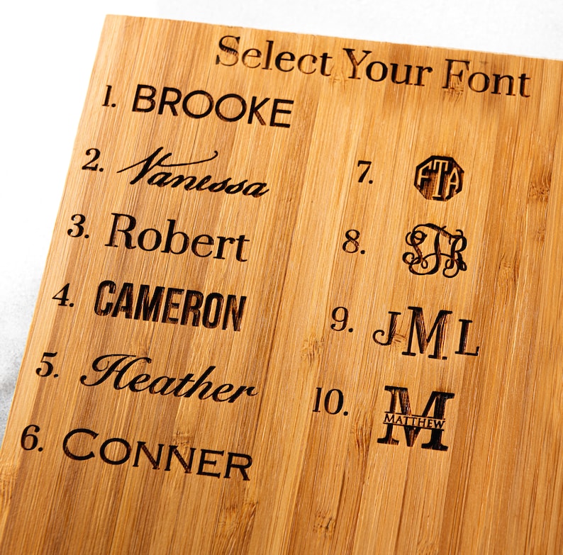 Op de afbeelding: Een houten snijplank met tien verschillende lettertypen om uit te kiezen voor een gepersonaliseerde gravure. De lettertypen zijn genummerd van 1 tot 10 en omvatten namen zoals Brooke, Vanessa, Robert, Cameron, Heather en Conner.