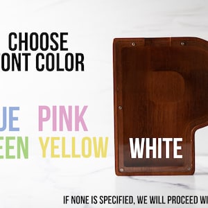 May include: A wooden piano-shaped box with a clear plastic window. The text "CHOOSE FONT COLOR" is above a list of color options: blue, pink, green, yellow, and white. The text "IF NONE IS SPECIFIED, WE WILL PROCEED WITH WHITE" is below the color options.