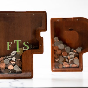 May include: Two wooden piggy banks shaped like the letters F and T. The F piggy bank has a clear plastic front and is filled with coins. The T piggy bank has a clear plastic side and is also filled with coins. The F piggy bank has the letters "FTS" written on the front.