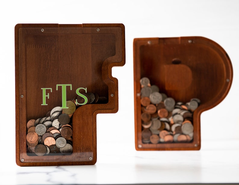 May include: Two wooden piggy banks shaped like the letters F and T. The F piggy bank has a clear plastic front and is filled with coins. The T piggy bank has a clear plastic side and is also filled with coins. The F piggy bank has the letters "FTS" written on the front.