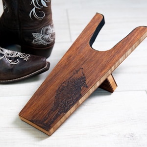 May include: A wooden boot jack with a wolf design. The boot jack is made of dark wood and has a black foam insert. The boot jack is on a white surface.