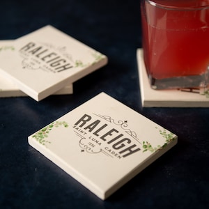 Puede incluir: Cuatro posavasos cuadrados blancos con diseños florales verdes y el texto "Raleigh Saint Luna Caden 1988" impreso en ellos. Un vaso de líquido rojo se encuentra sobre uno de los posavasos.