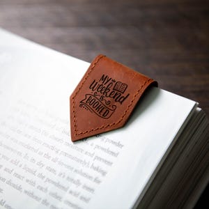 Op de afbeelding: Een bruine leren bladwijzer met de tekst "My Weekend is Booked" en een kleine illustratie van een boek.