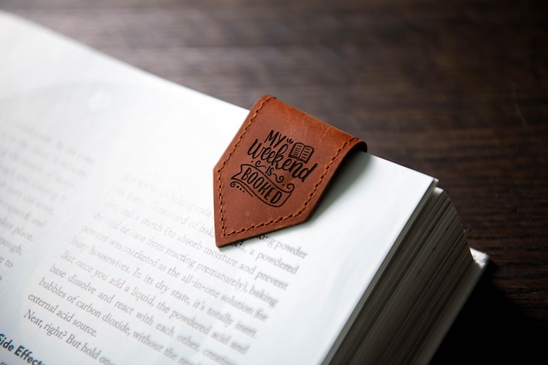 Op de afbeelding: Een bruine leren bladwijzer met de tekst "My Weekend is Booked" en een kleine illustratie van een boek.