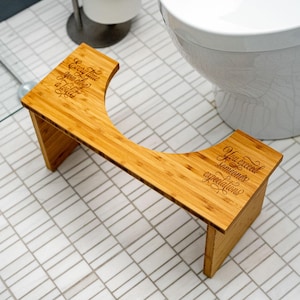 Peut inclure: Un tabouret de toilette en bambou avec un design incurvé. Le tabouret porte les inscriptions "Every time you use a toilet" et "You exceed someone's expectations". Fabriqué en bambou brun clair.