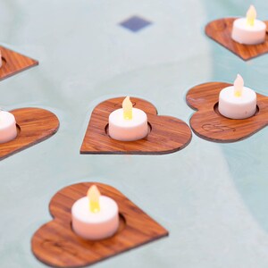 Op de afbeelding: Zes houten hartvormige kaarshouders met kleine witte theelichtjes die erin branden. De houders zijn gerangschikt op een lichtblauw oppervlak.