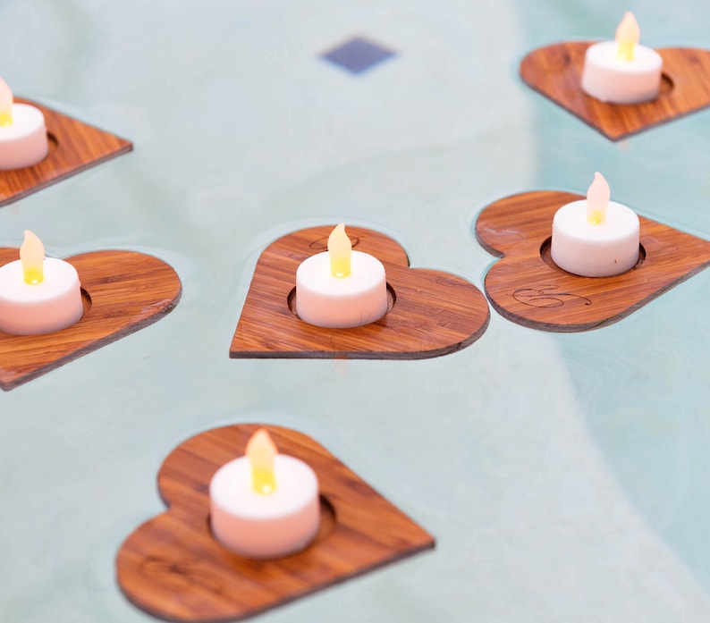 Op de afbeelding: Zes houten hartvormige kaarshouders met kleine witte theelichtjes die erin branden. De houders zijn gerangschikt op een lichtblauw oppervlak.