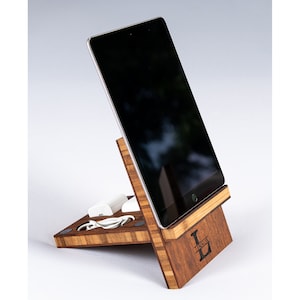Op de afbeelding: Een houten tabletstandaard met een gepersonaliseerde initial "L" en de naam "LESLIE" gegraveerd op de voorkant. De standaard heeft een gleuf voor oplaadkabels en een klein plankje voor een telefoon of ander apparaat. De standaard is gemaakt van licht en donker hout.