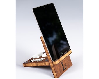 Op de afbeelding: Een houten tabletstandaard met een gepersonaliseerde initial "L" en de naam "LESLIE" gegraveerd op de voorkant. De standaard heeft een gleuf voor oplaadkabels en een klein plankje voor een telefoon of ander apparaat. De standaard is gemaakt van licht en donker hout.