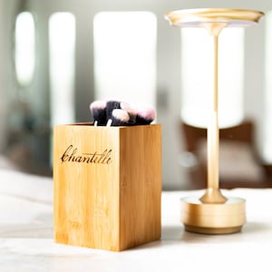 May include: A wooden makeup brush holder with the name "Chantelle" engraved on the front. The holder is filled with pink and black makeup brushes. The holder is sitting on a white surface next to a gold metal stand.