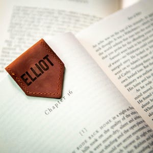 Op de afbeelding: Een bruine leren bladwijzer met de naam "ELLIOT" gegraveerd in zwarte letters. De bladwijzer heeft de vorm van een puntige driehoek en heeft een gestikte rand.