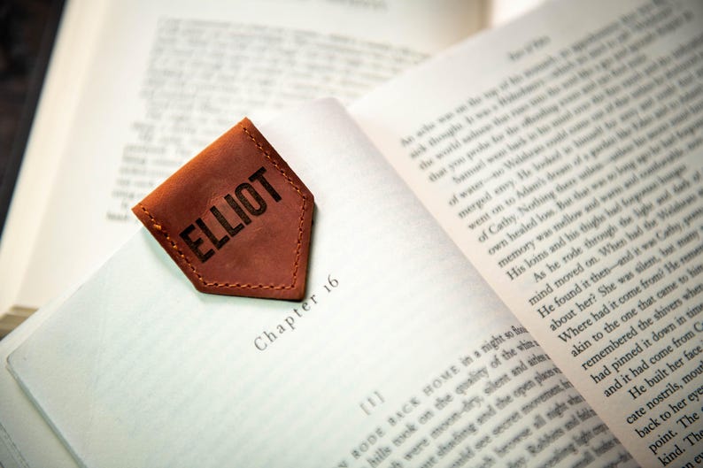 Op de afbeelding: Een bruine leren bladwijzer met de naam "ELLIOT" gegraveerd in zwarte letters. De bladwijzer heeft de vorm van een puntige driehoek en heeft een gestikte rand.