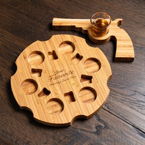 Puede incluir: Una bandeja de servir de madera con forma de rev&oacute;lver con un peque&ntilde;o vaso de chupito de vidrio en el mango. La bandeja tiene ocho hendiduras redondas para contener bebidas y el texto "Your Favorite Drinking Quote Here" est&aacute; grabado en la bandeja.