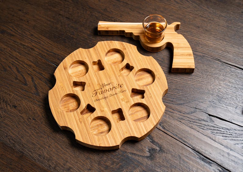 Puede incluir: Una bandeja de servir de madera con forma de rev&oacute;lver con un peque&ntilde;o vaso de chupito de vidrio en el mango. La bandeja tiene ocho hendiduras redondas para contener bebidas y el texto "Your Favorite Drinking Quote Here" est&aacute; grabado en la bandeja.