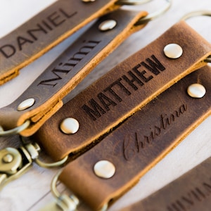 Pode incluir: Cinco chaveiros de couro castanho com argolas e fechos metálicos. Cada chaveiro é personalizado com um nome: Daniel, Mimi, Matthew, Christina e Johnny.
