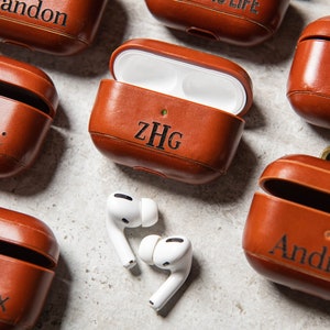 May include: Six brown leather AirPod cases with different names engraved on them. The names are: Brandon, Noemi, ZHG, QAL, Andrew, and Hendrix. Two white AirPod earbuds are lying on a white surface.