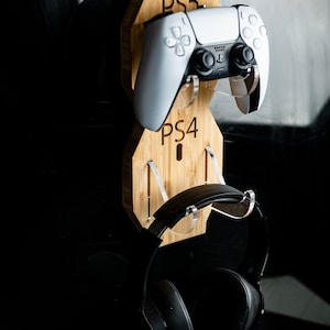 May include: A wooden gaming console stand with a white PS5 controller and black headphones. The stand is designed to hold a PS5 controller and a PS4 controller, with the text "PS5" and "PS4" visible.