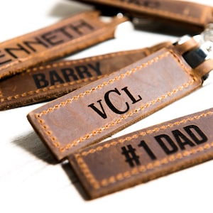 May include: Close-up of several brown leather keychains with silver key rings. Each keychain is rectangular and features stitched edges. The keychains are engraved with names and phrases, including "KENNETH", "BARRY", "VCL", and "#1 DAD".
