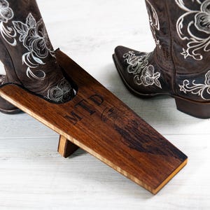 May include: A wooden boot jack with the initials "MTD" engraved on the side. A brown cowboy boot with white floral embroidery is being pulled off with the boot jack.