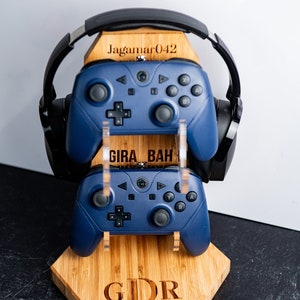 May include: A wooden stand with a personalized inscription "GDR" holds two blue video game controllers and a black headset. The stand also has the text "Jagamar042" and "GIRA BAH" engraved on it.