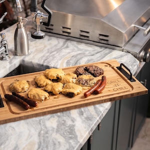 Puede incluir: Una tabla de cortar de madera con un mango negro y el texto "Grill N Chill" grabado en ella. La tabla está llena de comida a la parrilla, incluyendo hamburguesas, salchichas y otros artículos.