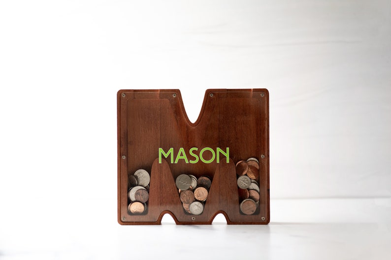 May include: Wooden coin bank shaped like the letter "M" with the name "MASON" printed on the front. The bank has three slots for coins, each filled with a small amount of change.