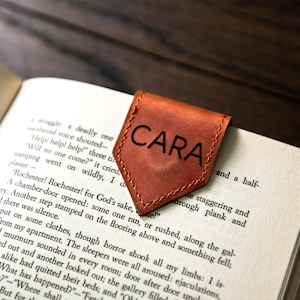 Op de afbeelding: Een bruine leren bladwijzer met de naam "CARA" erop gegraveerd. De bladwijzer ligt op een open boek.