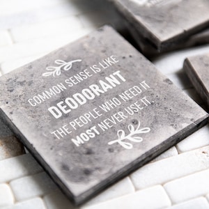 May include: Square, gray coasters with a textured appearance. The coasters feature white text that reads "COMMON SENSE IS LIKE DEODORANT THE PEOPLE WHO NEED IT MOST NEVER USE IT." Decorative leaf accents are also present.