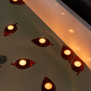 Op de afbeelding: Een wit bad gevuld met water en verlicht door drijvende kaarsen in blaadjesvormige houders. De kaarsen zijn in een patroon over het wateroppervlak gerangschikt.