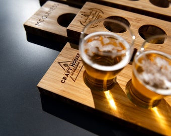 Puede incluir: Una bandeja de vuelo de cerveza de madera con el texto "CRAFT HOUSE" y "CRAFT BEER EXPERIENCE" grabado en ella. La bandeja tiene cuatro ranuras para vasos, y dos de las ranuras están llenas de vasos de cerveza.