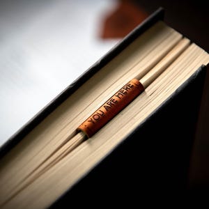 Op de afbeelding: Een bruine leren bladwijzer met de tekst "You are here" is geplaatst in een gesloten boek met een zwarte kaft.