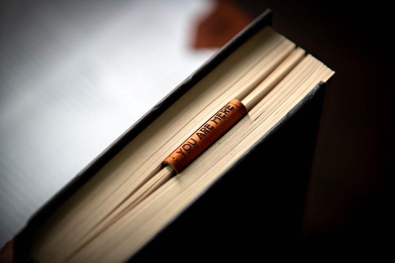Op de afbeelding: Een bruine leren bladwijzer met de tekst "You are here" is geplaatst in een gesloten boek met een zwarte kaft.