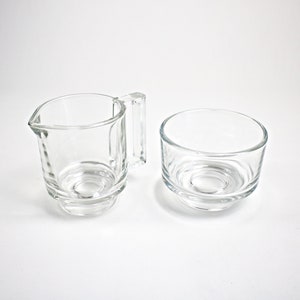 May include: A clear glass creamer and sugar bowl set. Both pieces have a simple, elegant design with a slightly flared base.
