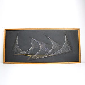 May include: Abstract string art on a black background, framed in wood. The artwork features three curved, sail-like shapes created with white and silver thread. The frame is a warm, natural wood tone, creating a striking contrast.