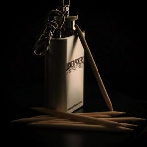 May include: A stainless steel toothpick holder with a keychain attachment, featuring the text "Licker Pickers Toothpicks". Several wooden toothpicks are scattered around the holder, with one leaning against it.