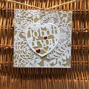 Può includere: Un biglietto bianco con un ritaglio a forma di cuore che dice "To both of you". Il cuore è decorato con glitter rosso e piccoli cuori rossi. Il biglietto è circondato da un motivo di pizzo bianco.