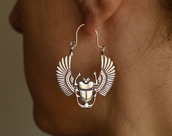 May include: Silver hoop earrings with a scarab beetle design. The beetle has wings and is detailed with intricate patterns.