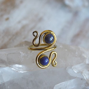 May include: A gold ring featuring two blue lapis lazuli stones set within the design. The ring is displayed on a white crystal background.