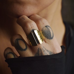 May include: A gold hammered ring with a split design. The ring is worn on the middle finger of a hand with tattoos.