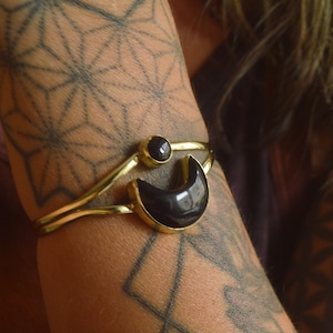 May include: A gold-toned metal cuff bracelet featuring a smooth, polished black crescent moon shape and a small, round black stone set above it. The bracelet has an open design with two thin bands that meet at the central black stone and moon.