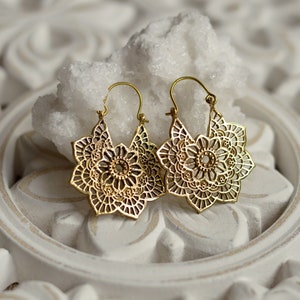 K&ouml;nnte beinhalten: Ein Paar goldfarbene, durchbrochene Ohrringe. Jeder Ohrring hat ein florales Design mit komplizierten Details. Die Ohrringe werden auf einer wei&szlig;en, verzierten Oberfl&auml;che mit einem wei&szlig;en Felsen dahinter pr&auml;sentiert.